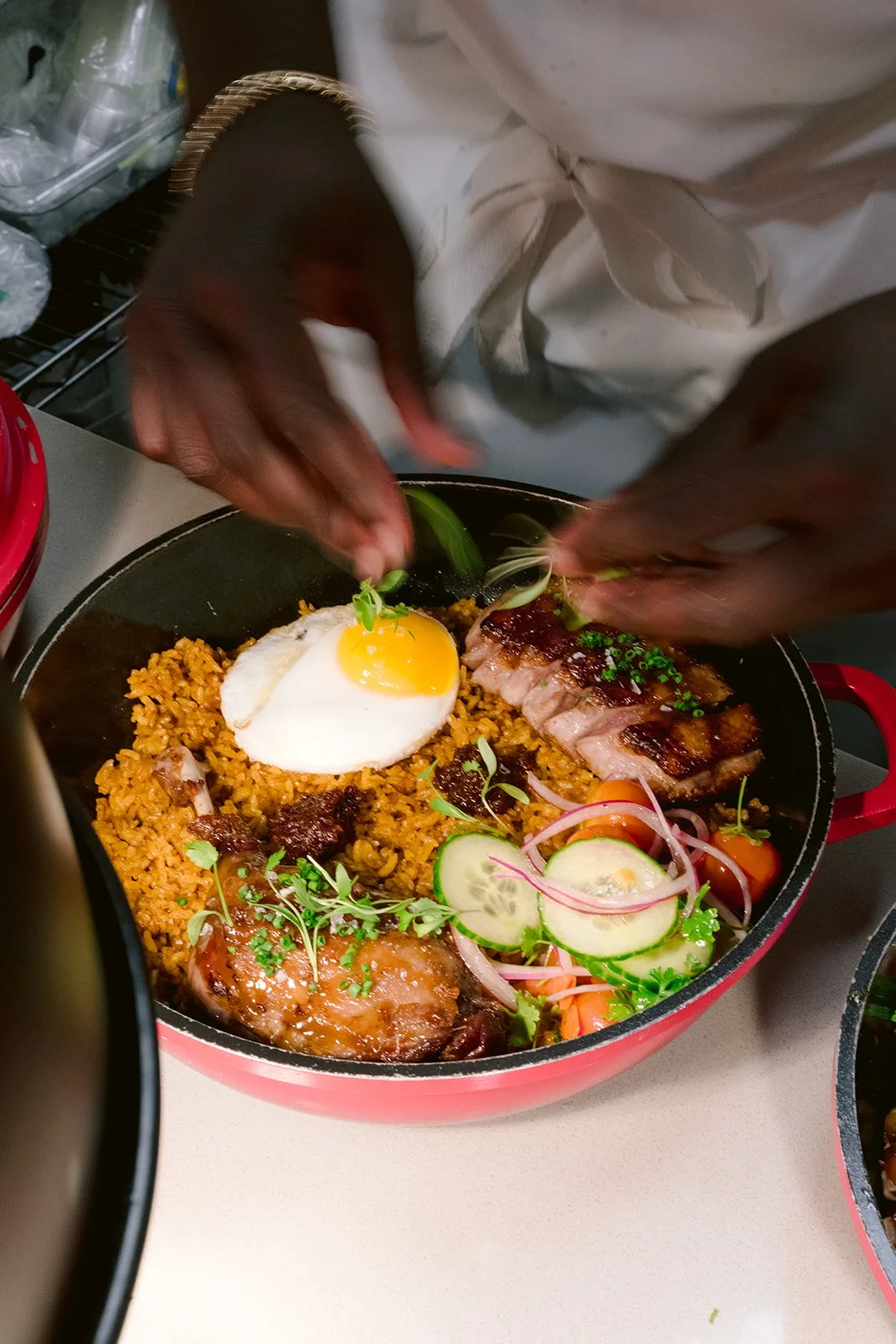 Chef Adjepong garnishing a bowl of spicy rice with a sunny side up egg, sliced cucumbers, cherry tomatoes, sliced onions, and grilled meat, with microgreens on top.